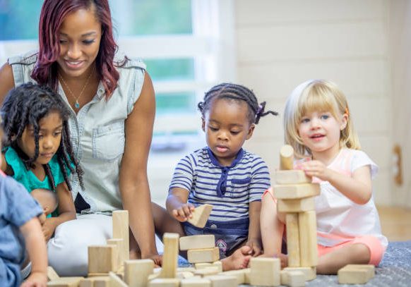 A preschool teacher of African descent is on the floor in a classroom with a group of children playing with wooden blocks.