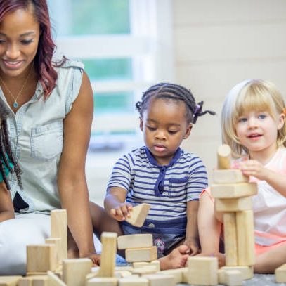 A preschool teacher of African descent is on the floor in a classroom with a group of children playing with wooden blocks.