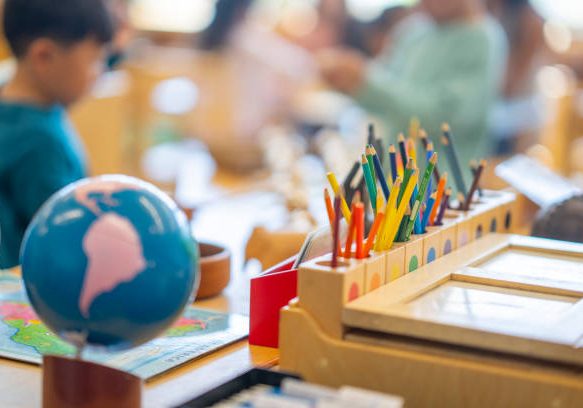 A Montessori Geography classroom set-up complete with a world map, globes and pencil crayons all sitting out on a desk.  Two young students can be seen blurred in the background working away.