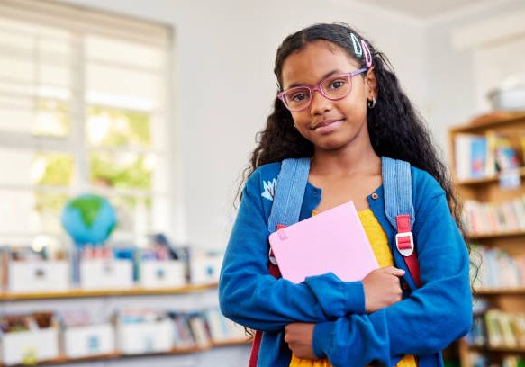 Portrait of confident black girl wearing backpack and holding agenda while looking at camera. Cheerful indian girl wearing eyeglasses and standing in classroom with copy space. Happy smiling middle eastern schoolgirl standing in primary class, back to school.
